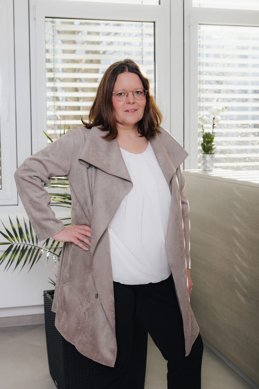 Frau in beiger Jacke steht lächelnd im Büro.