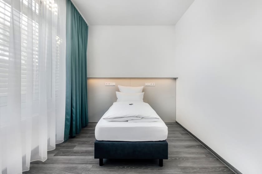 Helles Hotelzimmer mit Einzelbett und Vorhängen.