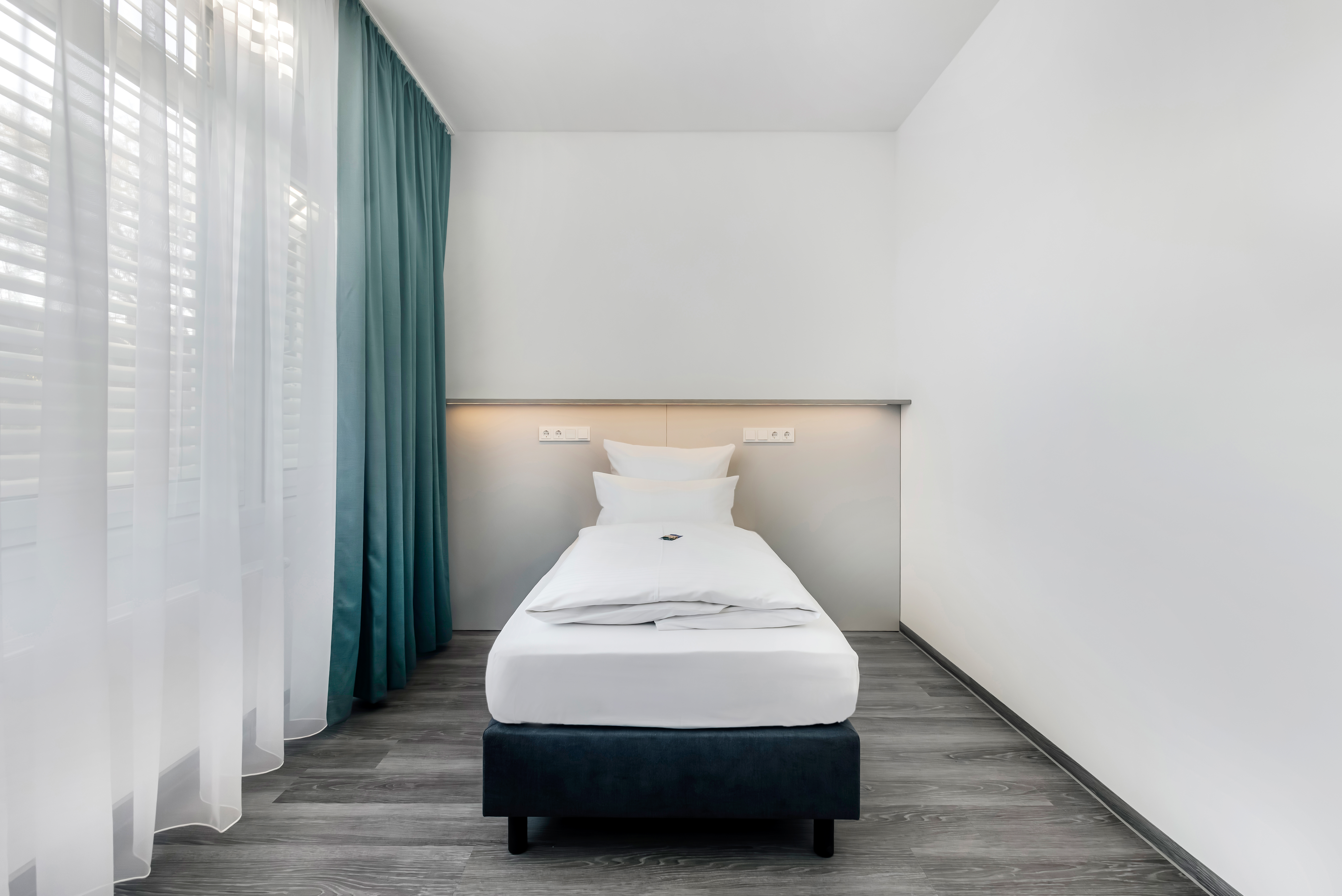 Helles Hotelzimmer mit Einzelbett und Vorhängen.